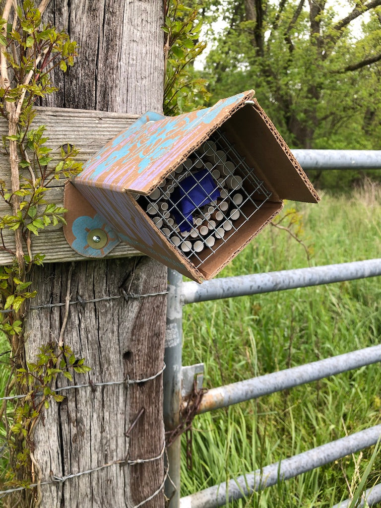 A bee hotel secured to a gate near a field.